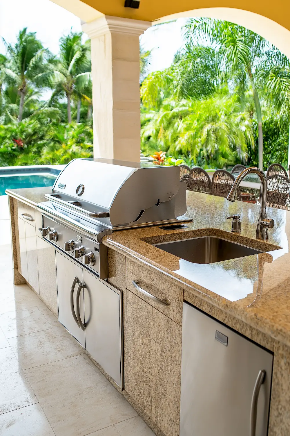 Outdoor kitchen with stainless steel grill and sink surrounded by palm trees and a pool